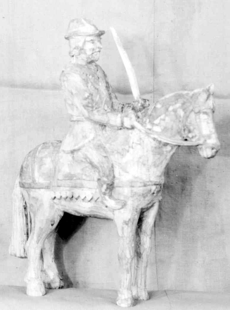 Figur av tre, forestiller hest med rytter. Mannen har hatt og sverd i hånden, og hestens seletøy i den andre hånden.