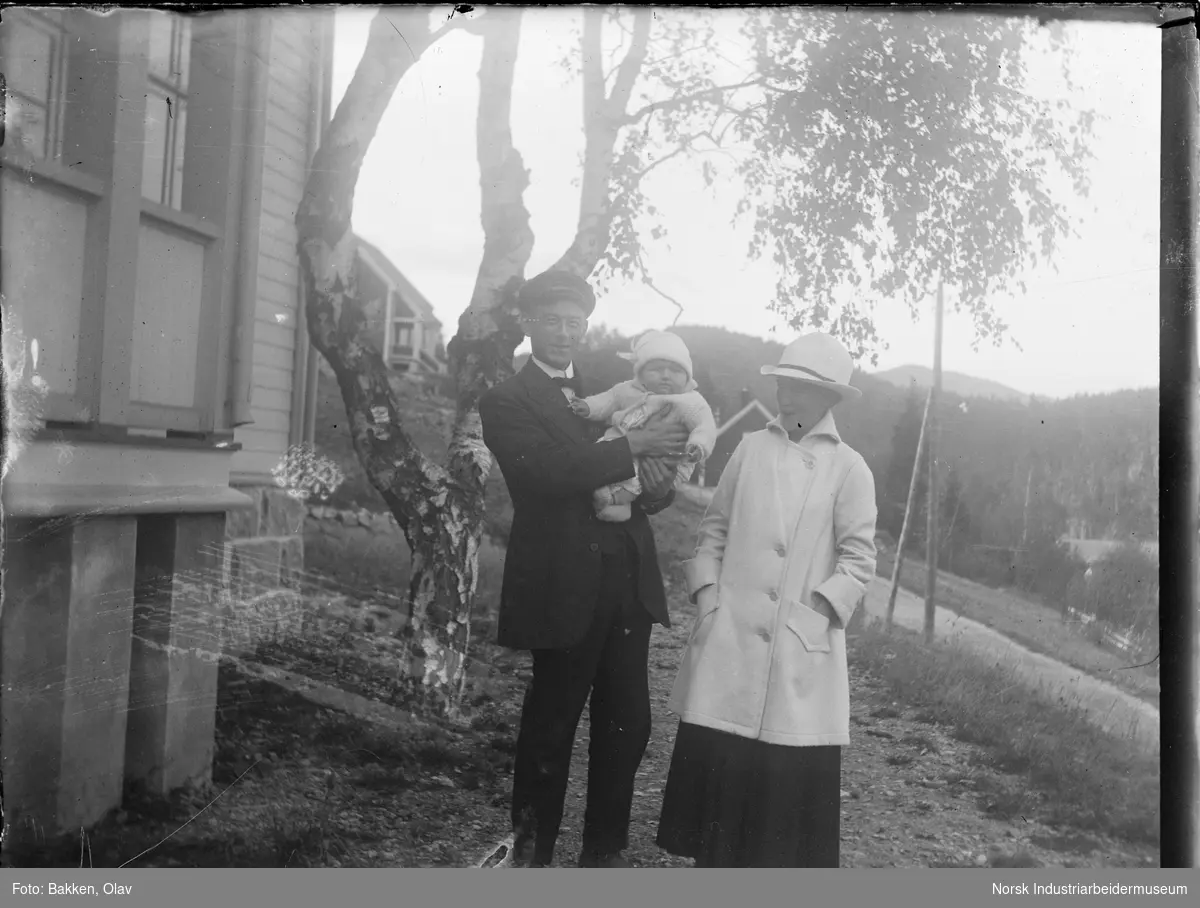 Kvinne og mann stående foran bolighus, mannen med spedbarn på armen.