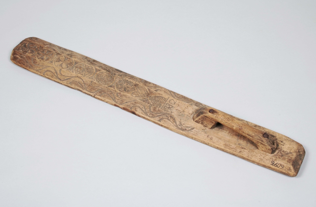 Mangelbräde i brunbetsat trä (färgslitage). Påspikat handtag på ena änden. Brädet med upphöjt mittparti och avfasade kanter. Rundad framtill. Längs mitten, skuren dekor av hjärtan, bladrosetter virvelhjul m.m. samt årtalet "1782". Längs kanterna stiliserad bladranka.

Mangelbräde eller kavelbräde är ett redskap av trä som använts för mangling av textilier. Mangelbrädet användes tillsammans med en kavel, en slät rulle av trä. Den textil som skulle manglas, rullades upp på kaveln, på ett bord eller annat slätt underlag. Under hårt tryck, rullades kaveln med hjälp av mangelbrädet, fram och tillbaka över bordet. (Wikipedia)