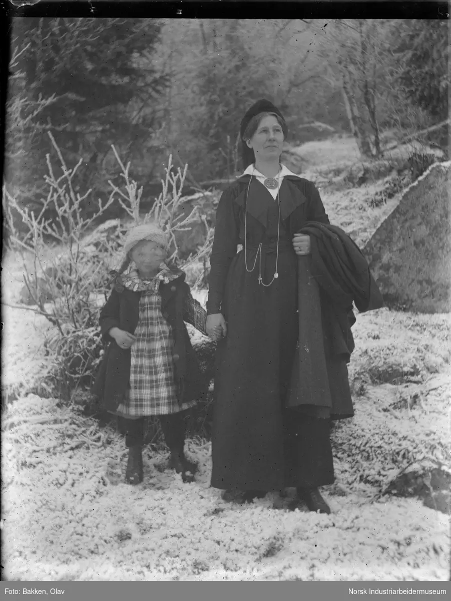 Kvinne holder jente i hånden oppstilt utendørs i snø. Kvinnen har vinterkåpe hengende over armen, jenta har vinterkåpen på seg.