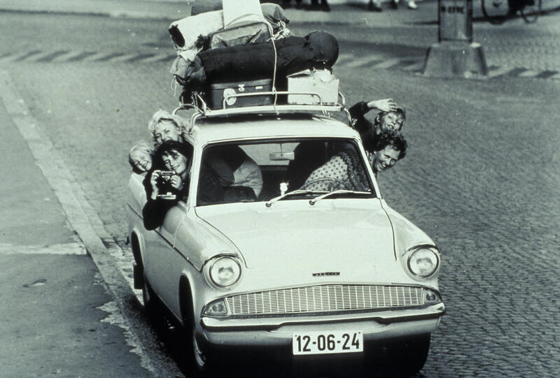 Fotografi av bil med bagasje på taket. Fem personer stikker hoet ut av vinduene i bilen.