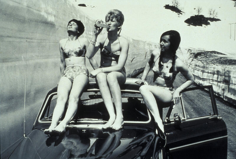 Fotografi av tre unge demer i bikini som sitter på taket av en bil. En drikker fra en glassflaske. Rundt dem er det snø.