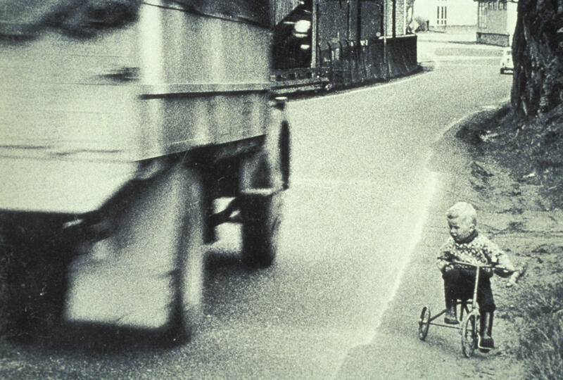 Fotografi av et lite barn på trehjulssykkel langs en vei med tung trafikk.