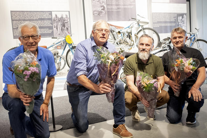 Fire menn sitter på kne med hver sin blomsterbukett foran utstillingen Basse i 100.