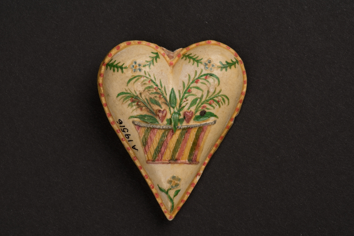Kotiljongsmärke, troligen av papier-maché, i form av ett stoppat hjärta.
Hjärtat är dekorerat på bägge sidorna och har en kant målad i gult och rött. På den ena sidan avbildas en randig blomsterkorg som innehåller gröna blad och röda hjärtan under en girlang. På andra sidan är en palm under girlang. På denna sida finns även en inskrift i blyerts: Söderköping 1850.