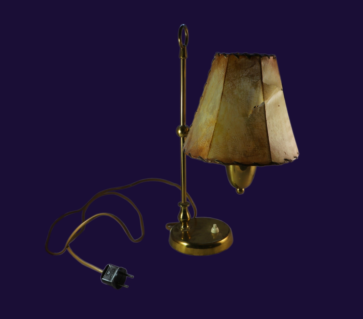 Elektrisk lampa i mässing. Rund, platt fot med lampknapp. En stång med en rund ring högst upp. I mitten på stången är en kula där det går ut en stång som själva lampan sitter på. Lampdelens underdel är kupformad. Lampskärm i pergament på metallställning.

Tillstånd vid förvärv: Lampskärmen är sprucken.,