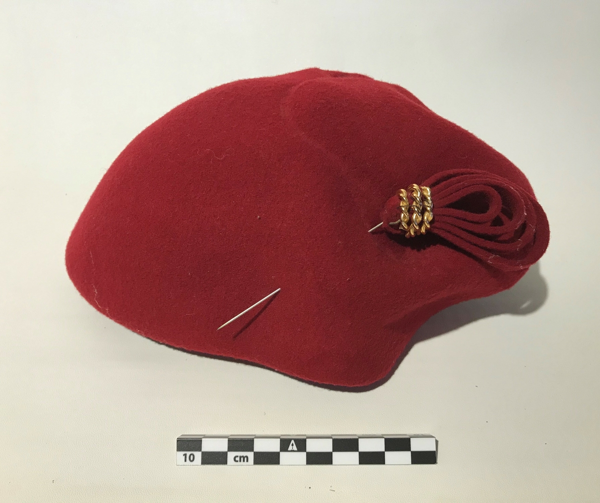 Hatt. Rød damehatt i ullfilt med en stor pin til pynt. Hatten har strikk til å ha under haken.