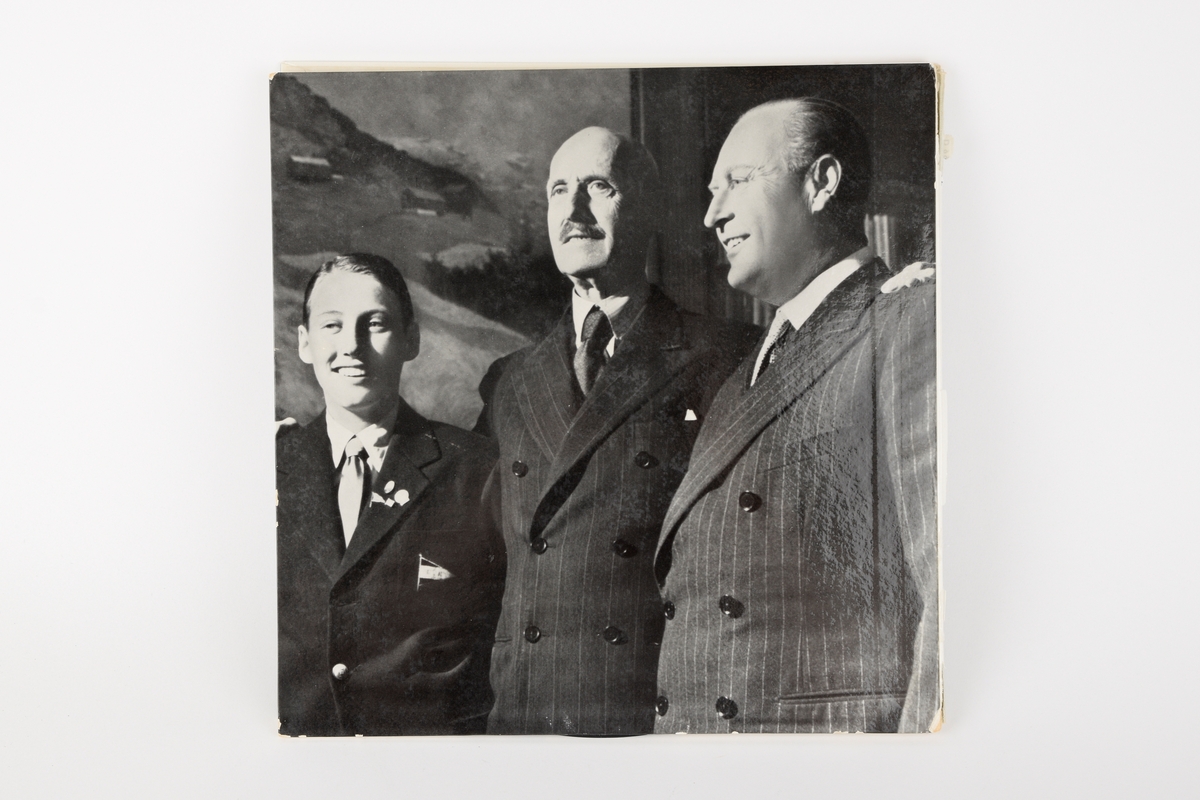 På fremsiden: Svart-hvitt foto av Kong Haakon VII sittende i en stol med ei åpen bok i fanget. På baksiden: Svart-hvitt foto av prins Harald, kong Haakon og kronprins Olav, tatt på Skaugum i 1952.