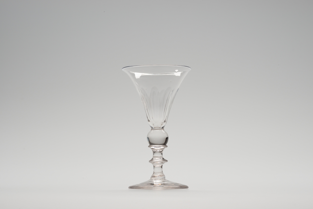 Stetteglass med rund fot og profilert stett. Cupa sterkt utsvunget ved munningen. Lang-ovale slipninger langs glasset.
