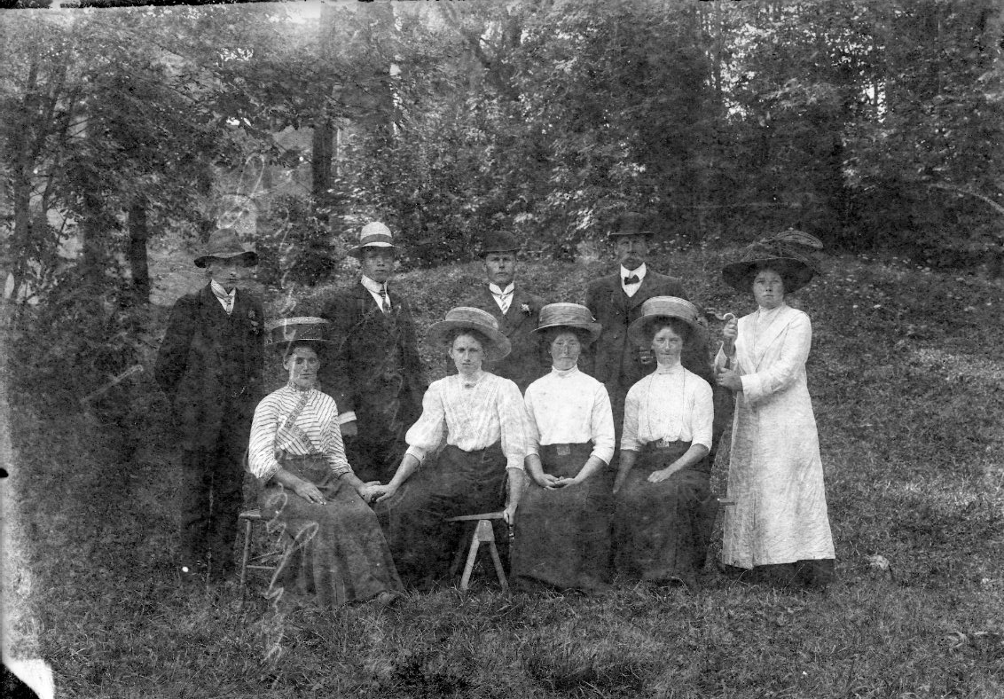 Fyra män och fem kvinnor, alla med hatt, står och sitter på en grässlänt vid Visingsborgs slottsruin. Det är sommar.