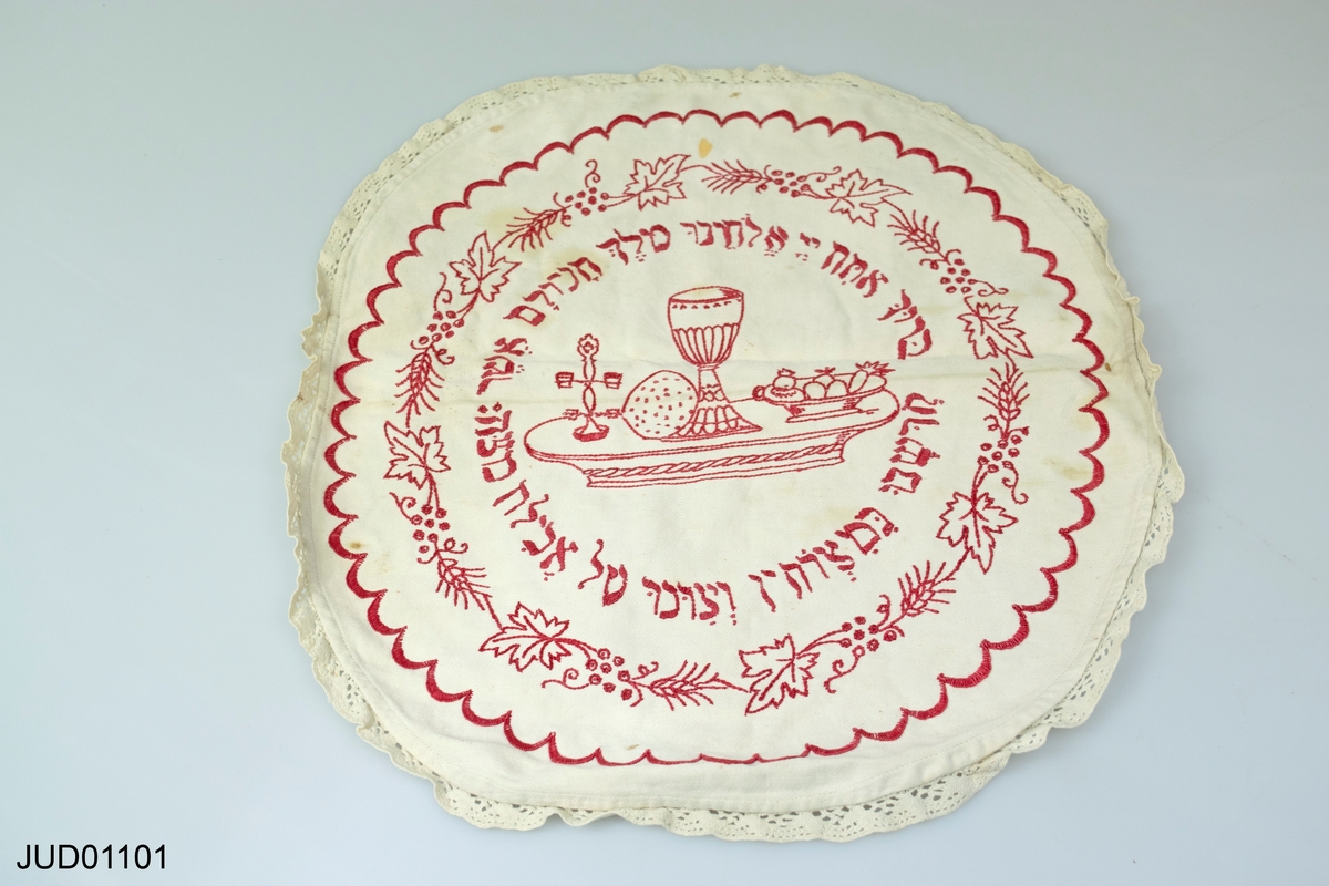 Rund duk på vit botten med röd brodyr. Motiv av pesachmåltiden och välsignelse på hebreiska för ätandet av matsabrödet runt om.