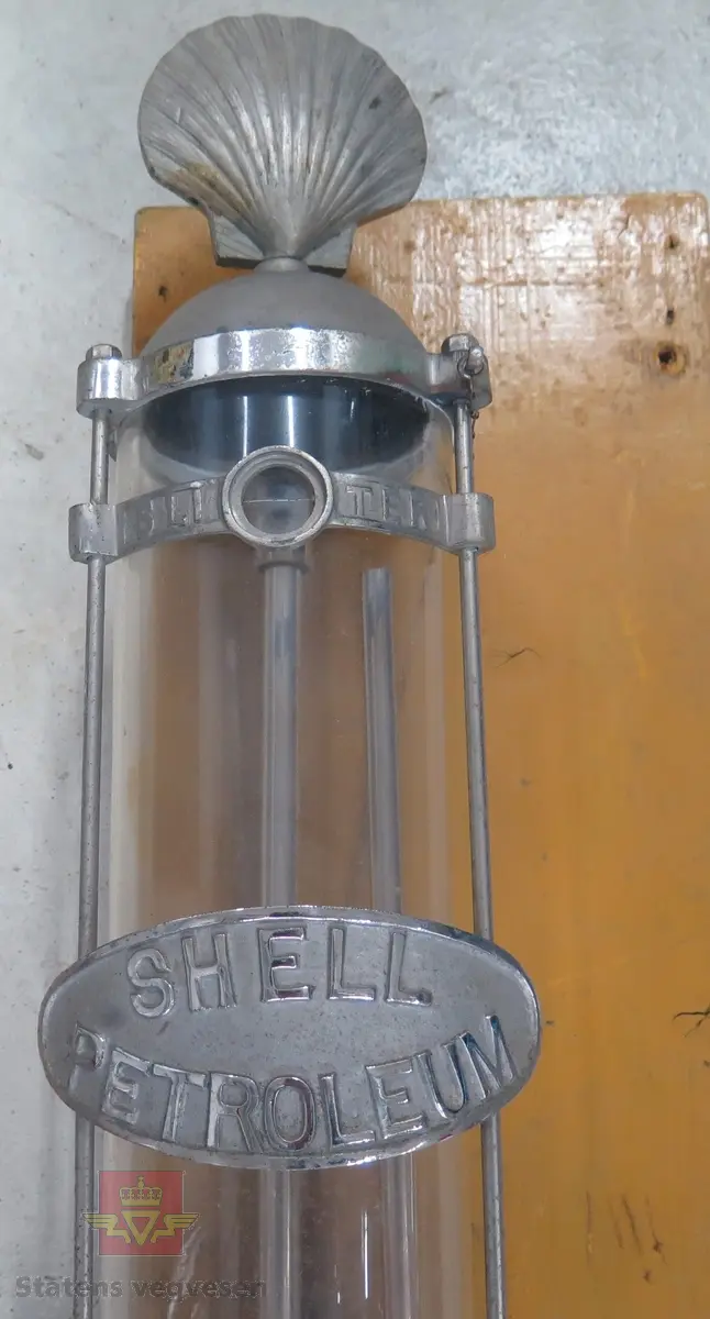 Hånddrevet parafinpumpe på treplate. Har en rund glassbeholder med en Shell-logo på toppen. Beholderen er påkoblet en manuell pumpe, hvor et metallrør skal gå videre ned i parafintønnen.