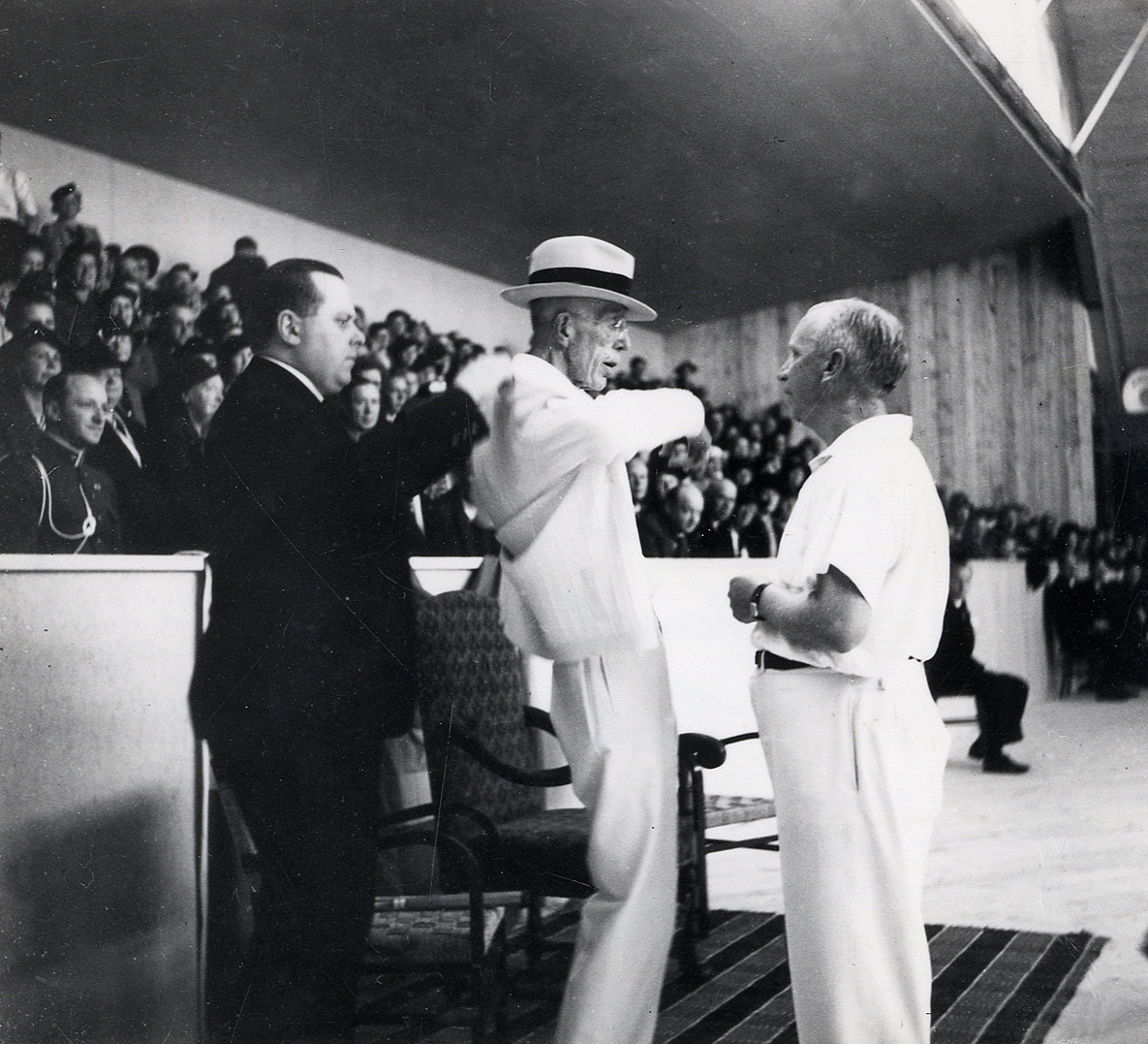 Kung Gustaf V vid invigningen av tennishallen (idrottshallen) i Växjö 1938. Kungen samtalar med dåv. läroverksadjunkt Thure Fridlund, samtidigt som han får hjälp med sin kavaj.