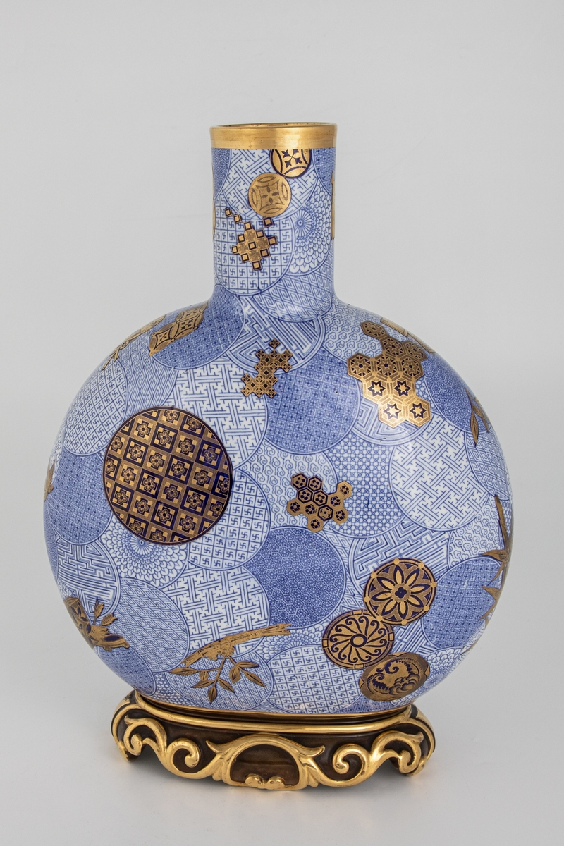 Stor vase med sylinderformet hals og flattrykt kuleformet korpus på kraftig fot med forgylt rankedekor. Mønstret sirkeldekor i blå underglasur, samt bambus, kostbare gjenstander og sirkelmønstre i mørkere blått og gull.