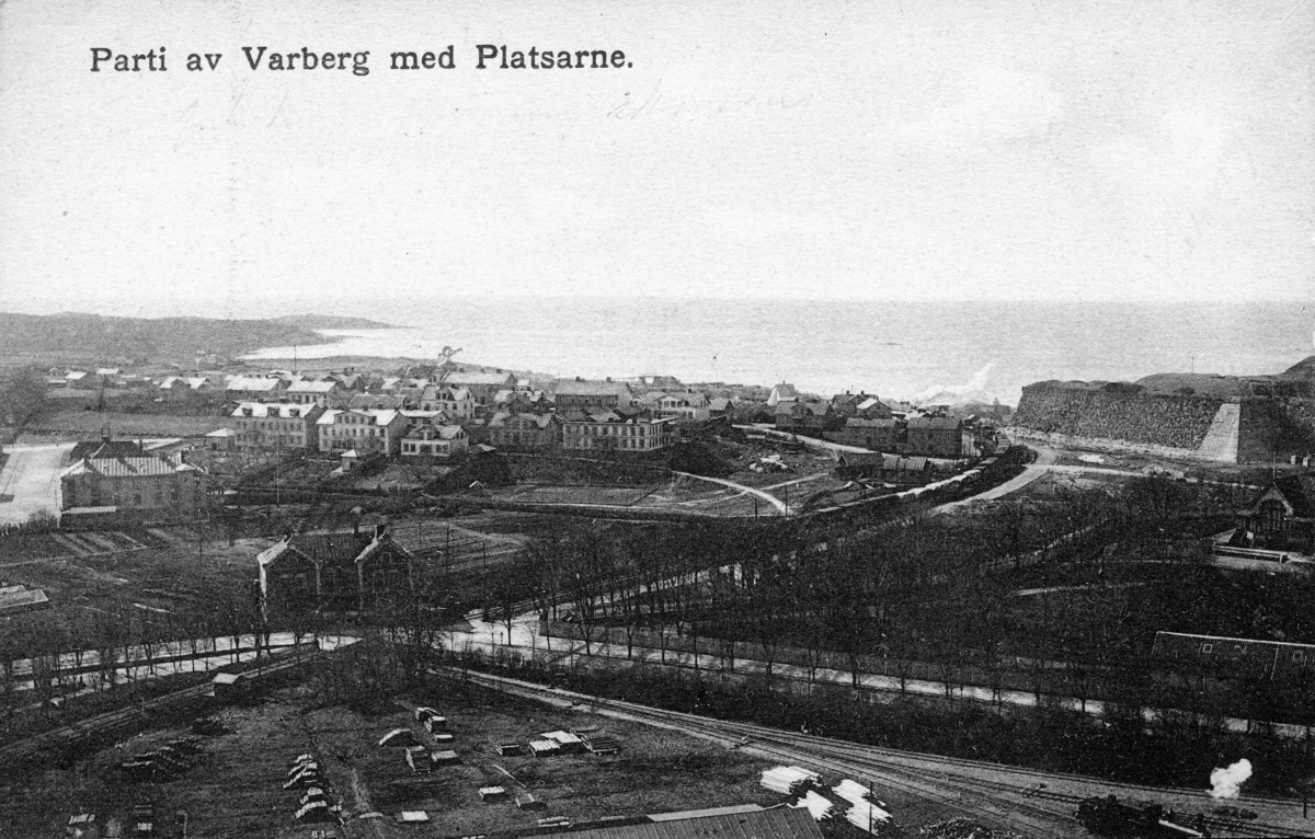 Vykort: "Parti av Varberg med Platsarne". Utsikt från kraftstationens skorsten mot Platsarna. Ett tåg är på väg söderut - ångan från loket syns vid södra fästningshörnan. Till höger i bild syns societetsparken med restaurangen. I förgrunden upplag av bland annat brädor. En järnväg söderut från Varberg anlades under namnet Mellersta Hallands järnväg och invigdes 1886.