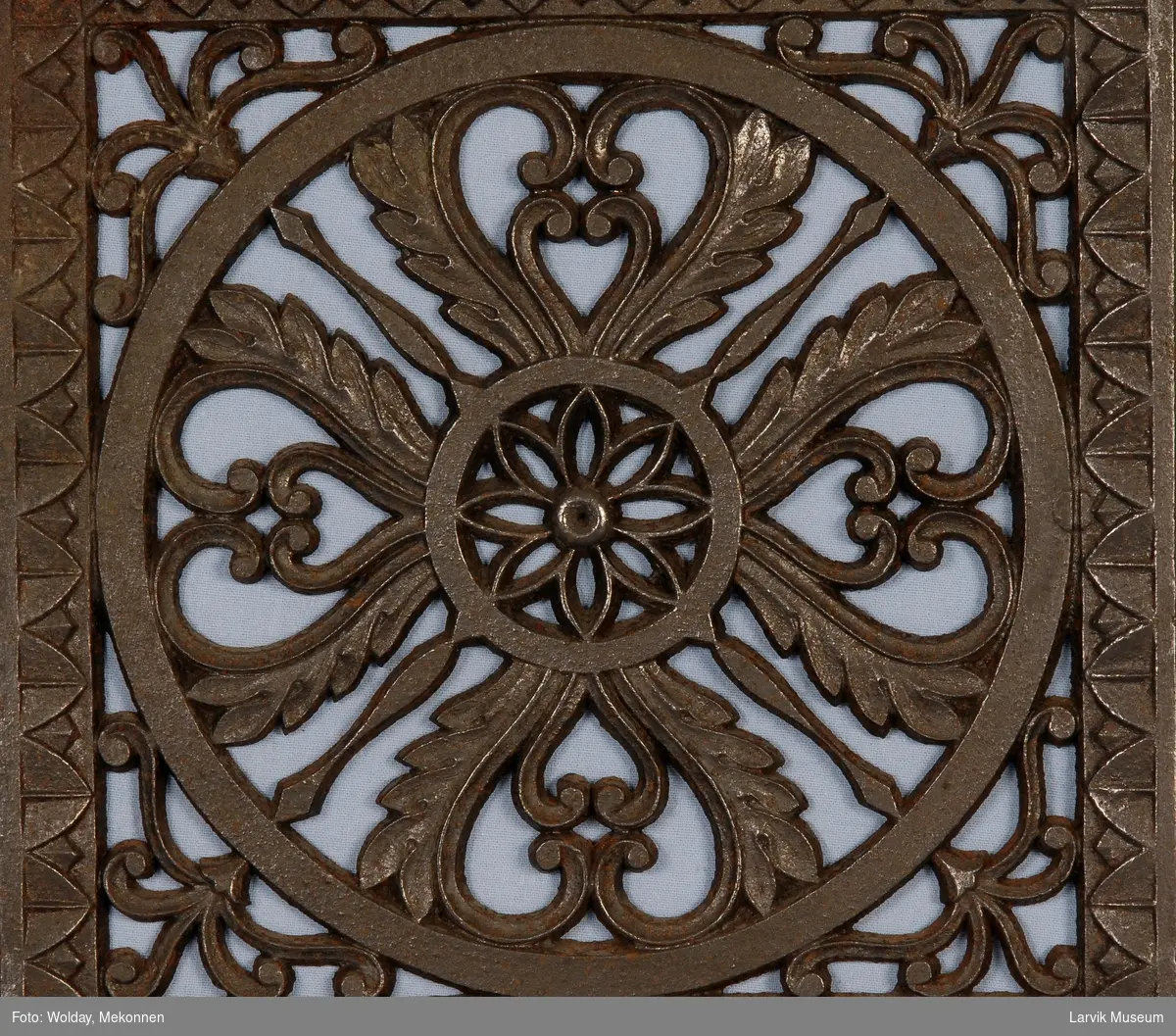 Symetrisk gjennombrudt blomster-ornamentikk i en sirkel, ornamentikk i hjørnene avsluttes med en bord-ramme
