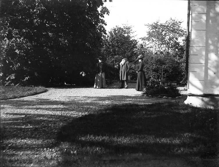 Två kvinnor och en man med långt ljust skägg står på en gårdplan framför en knut. Till höger en putsat husknut.