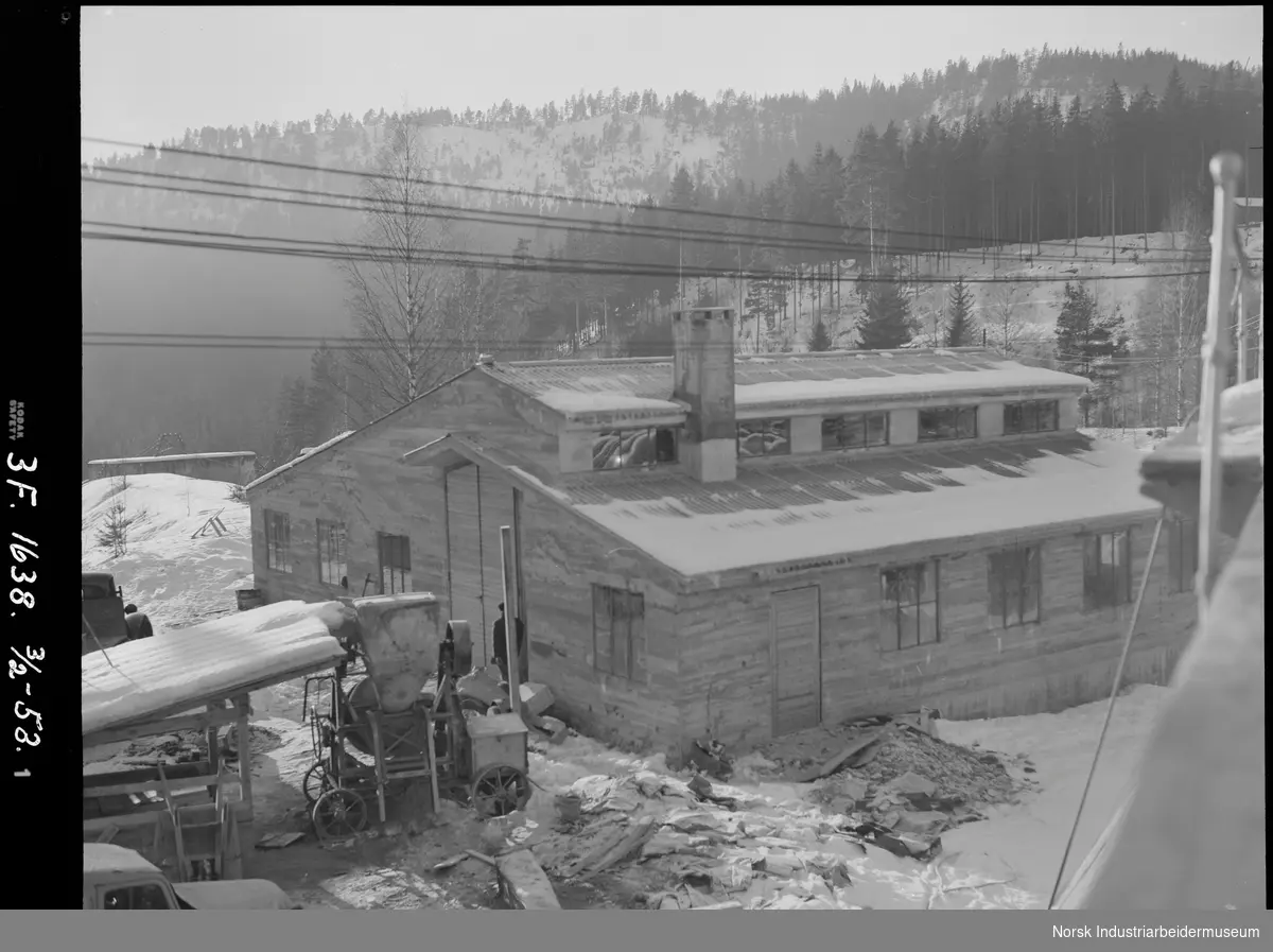 Svælgfos kraftstasjon (Svelgfoss kraftstasjon). Arbeid med nytt verksted. Mann stående ved vegg på bygg. Maskin som frakter masse på forsiden av bygg. Trillebår og lastebiler oppstilt rundt.