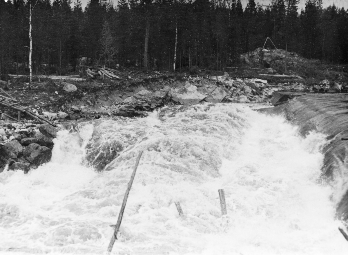 Tømmerfløting i elva Søndre Osa i Åmot kommune i Hedmark. Fotografiet er tatt våren 1936, ved den da nybygde kraftverksdammen ved Kvernfallet. I forgrunnen ser vi hvordan vannet og tømmerstokkene fløt over den avrundete damkrona. Noen av stokkene reiste seg i strømkavet under dammen, før de fløt videre. Den underliggende konstruksjonen, som vi ikke ser på dette bildet, var utført i armert betong. Fallhøyden var heller ikke så stor at de stokkene som landet med ei av endeflatene under dammen ble påført skader. Kvernfalldammen kom ikke til å skape nevneverdige problemer for tømmerfløtinga. Til venstre i bildet skimter vi den nedre delen av et trestillas, et skråplan med skinnegang for vogner som antakelig hadde vært brukt i anleggsperioden. Til høyre i bakgrunnen sto det en stubbebryter.