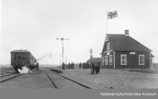 Grimetons järnvägsstation, som uppfördes vid Varberg-Ätrans järnväg år 1911. Ett tåg står och ångar på ena spåret och på stationsplanen finns en skara människor.