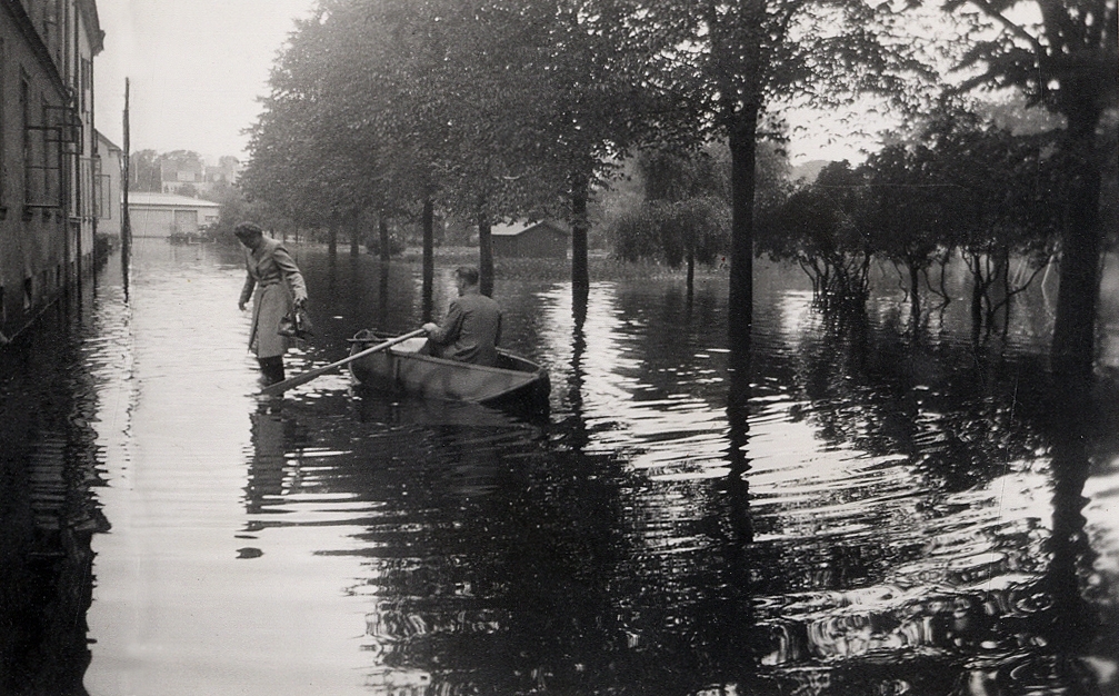 Översvämning på Biblioteksgatan, i riktning mot Liedbergsgatan, Växjö, 1940. 
En kvinna som är på väg in in ett bostadshus, passeras av en man i en roddbåt.