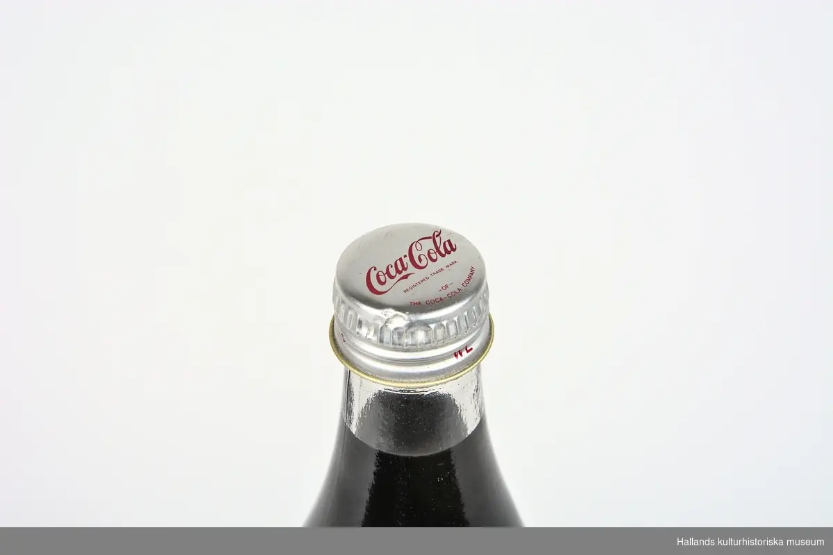 Läskedrycksflaska: Coca-Cola. Flaskan är tillverkad av klart glas och har en lång och sirlig flaskhals. 710ml. Oöppnad.