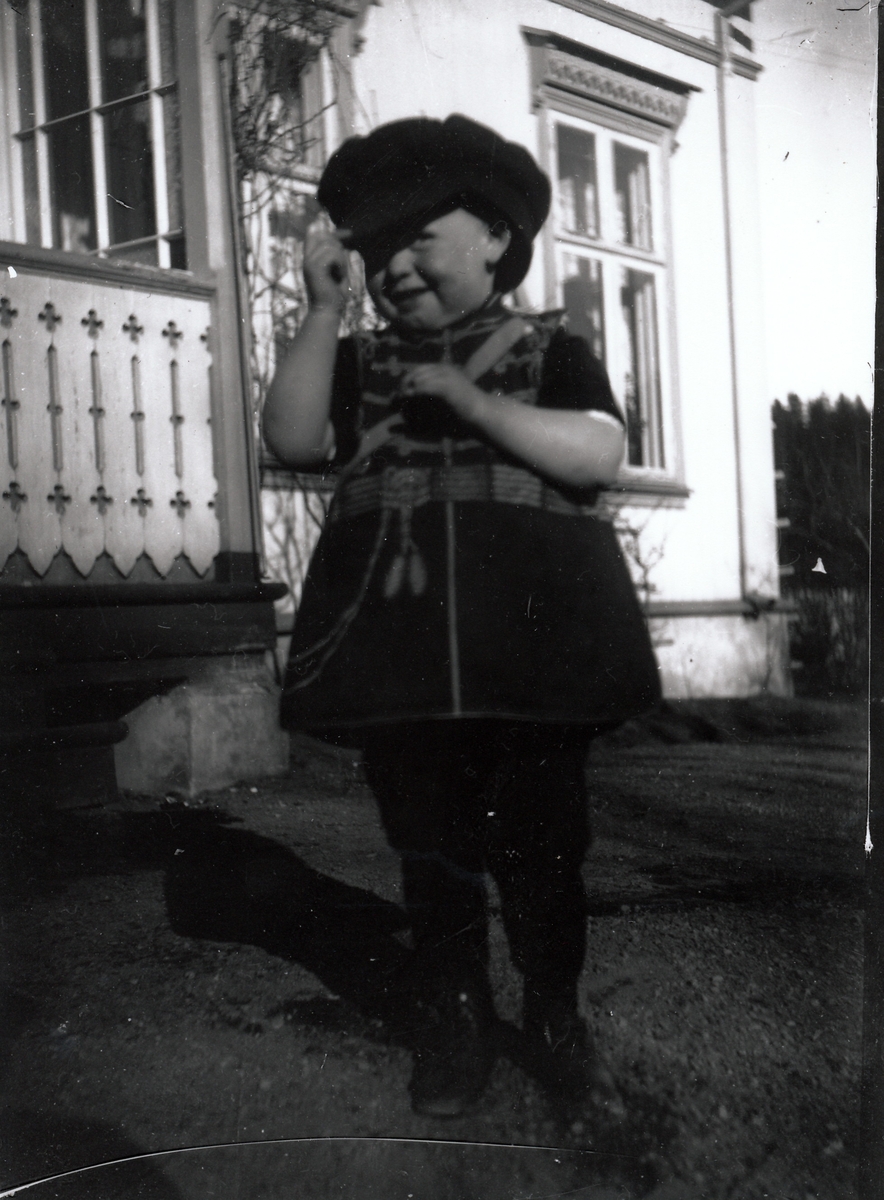 Liten gutt på ca. 2 år iført stor skyggelue og vest med mønster som ligner militæruniform. Står foran villa med sveitser stilpreg.