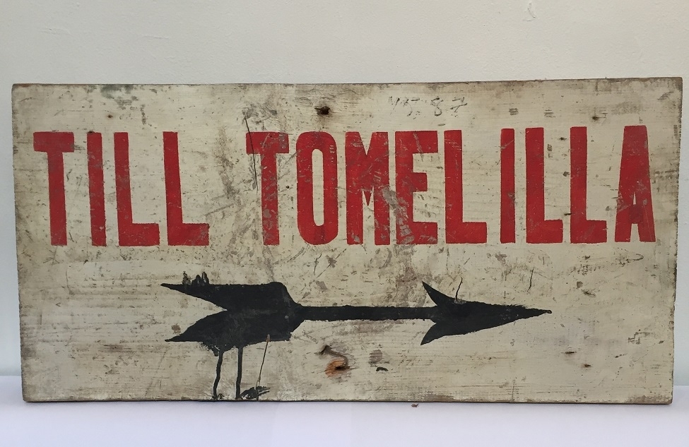 Träskylt, vit botten målad röd text "TILL TOMELILLA" med pil åt höger under texten, svartmålad.