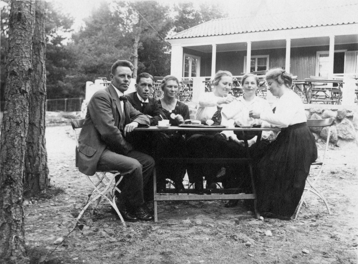 Två män och fyra kvinnor sitter runt ett bord utanför cafét i Folkets Park i Linköping. 1920-30-tal. En kvinna häller upp ur en kaffekanna.