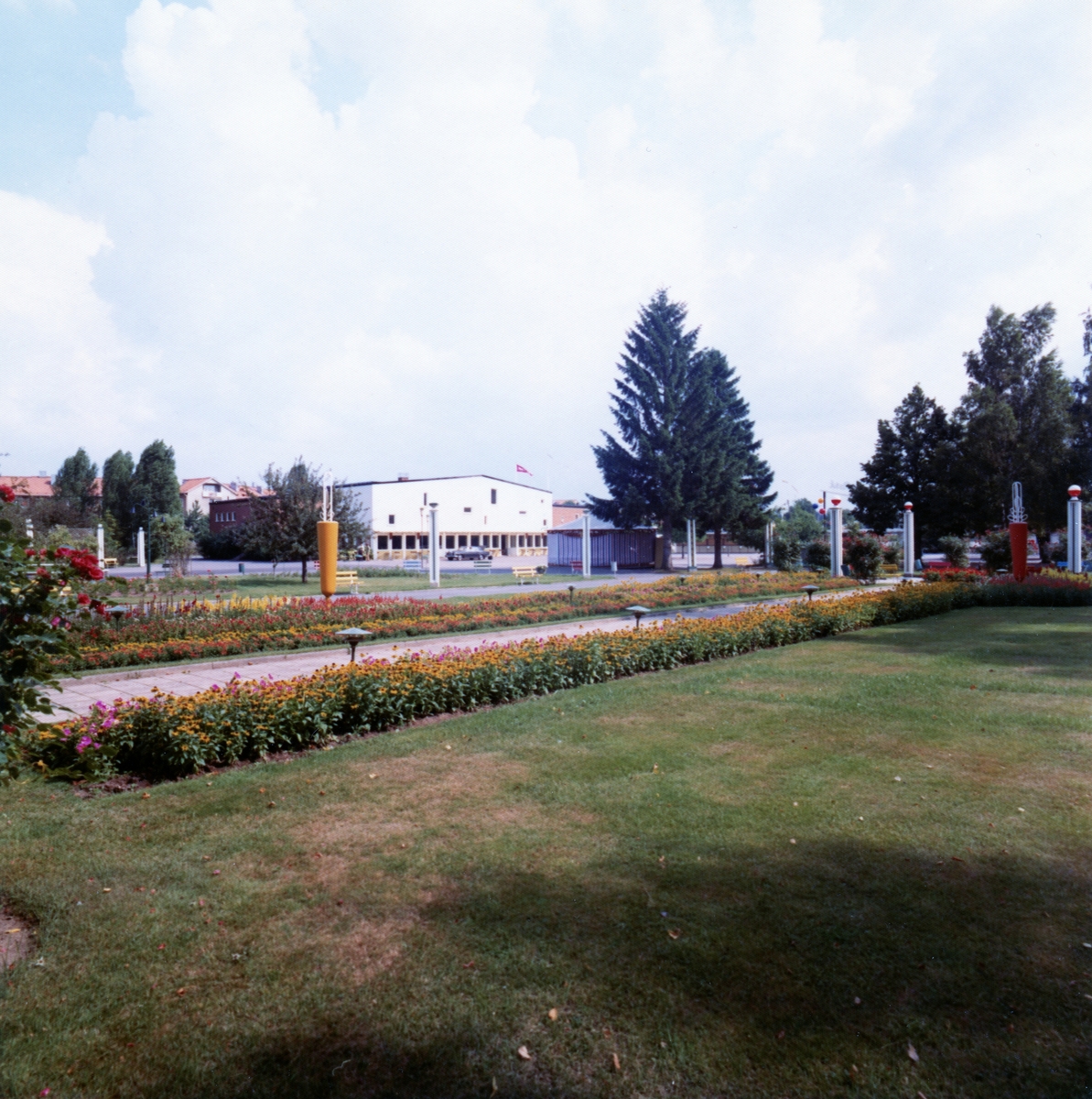 Bild från parken i Folkets Park. I bakgrunden en stor vit byggnad. I förgrunden en gräsmatta och en blomrabatt längs med gången.