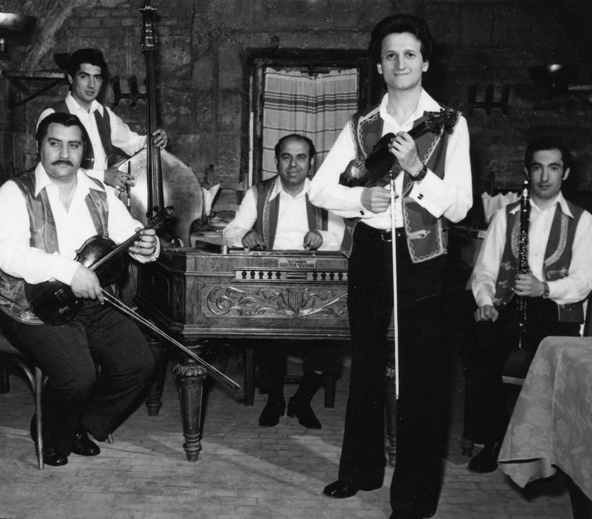 Svartvitt fot av fem män med instrument. Fyra sitter och en man står i förgrunden med sin fiol.
