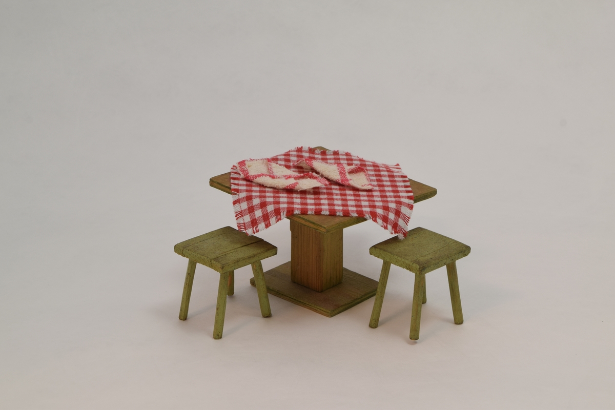 Kjøkkenbord med to krakker i tre. På bordet ligger det en rødrutete duk og to servietter. Møblene er laget av polske krigsfanger og tekstilene er tilført av tidligere eier.