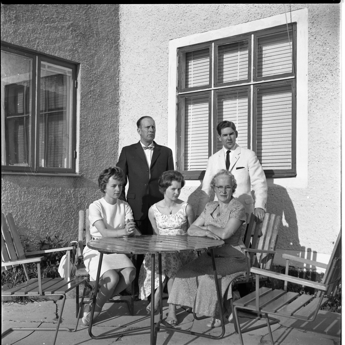 Fem finklädda personer befinner sig runt en trädgårdsmöbel, tre sitter och fyra står. Solen skiner på dem.