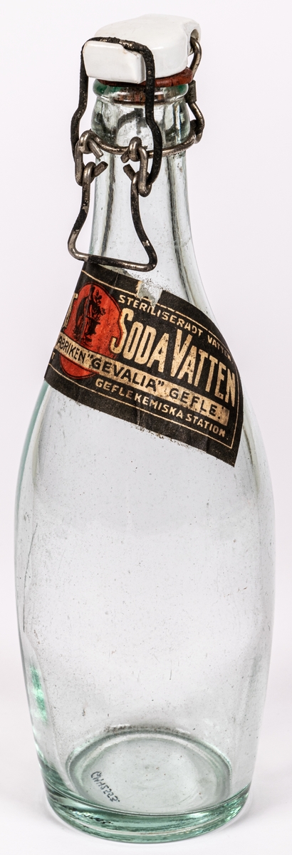 Flaska av genomskinligt glas, bukig form. Patentkort av keramik. Klistrad pappersetikett: Engelskt sodavatten, Mineralvattenfabriken Gevalia, Gefle