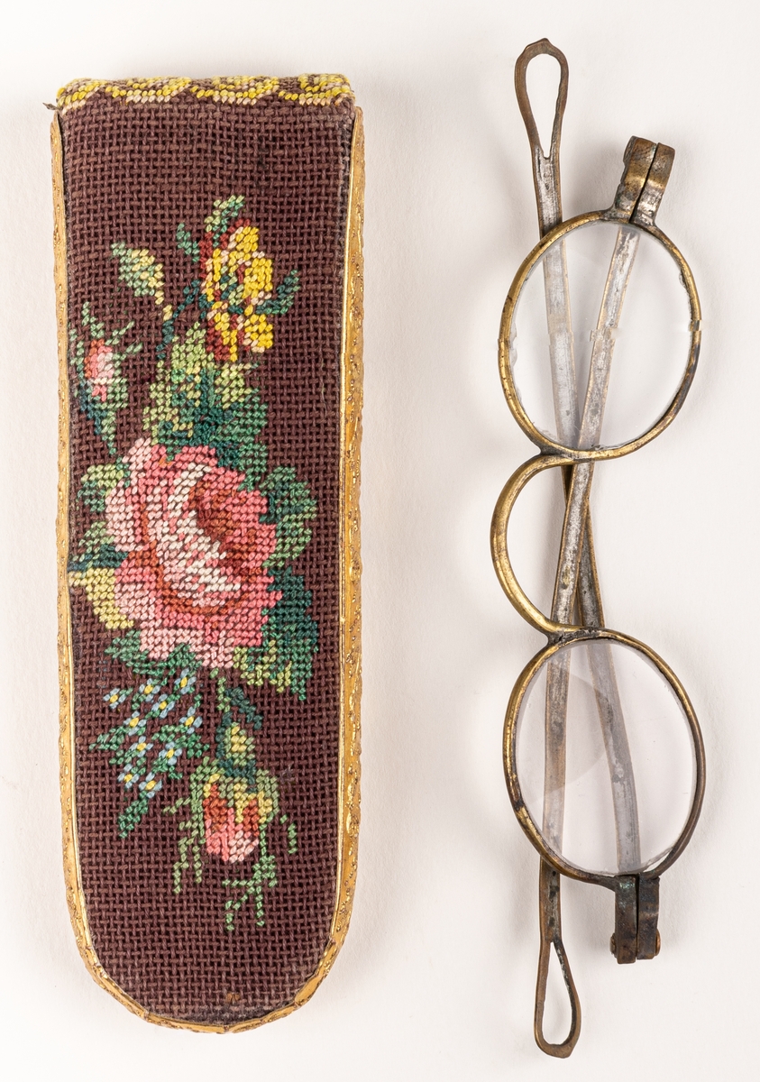Glasögonfodral, broderat, avlångt platt, nertill ruindat, av brunt linne med broderade blomster och "MT". Guldpapper med pressade ornament på smalsidorna.