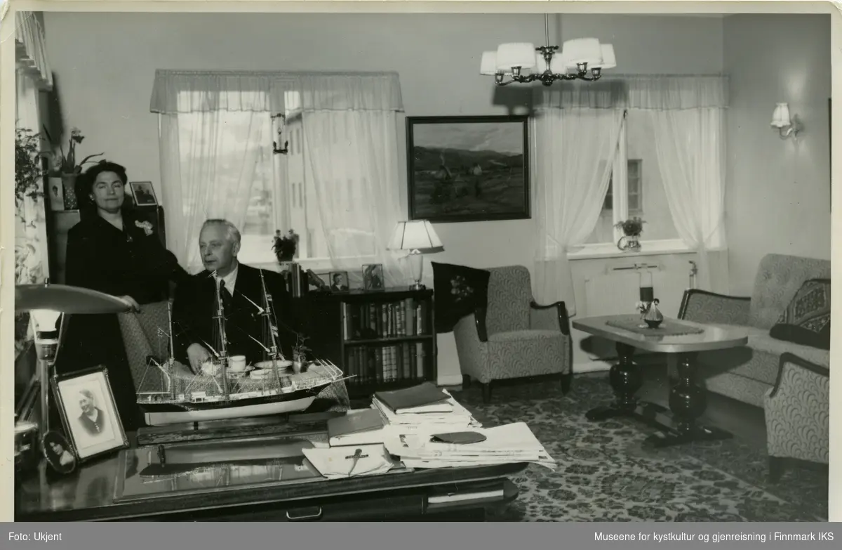 En man sitter i en lenstol bak et skrivebord. På skrivebordet står det en modell av en tremaster. En kvinne står bak han. Rommet har tre vinduer og er innredet med tidstypisk interiør fra 1950-tallet. Det er instalert fjernfarme i boligen.