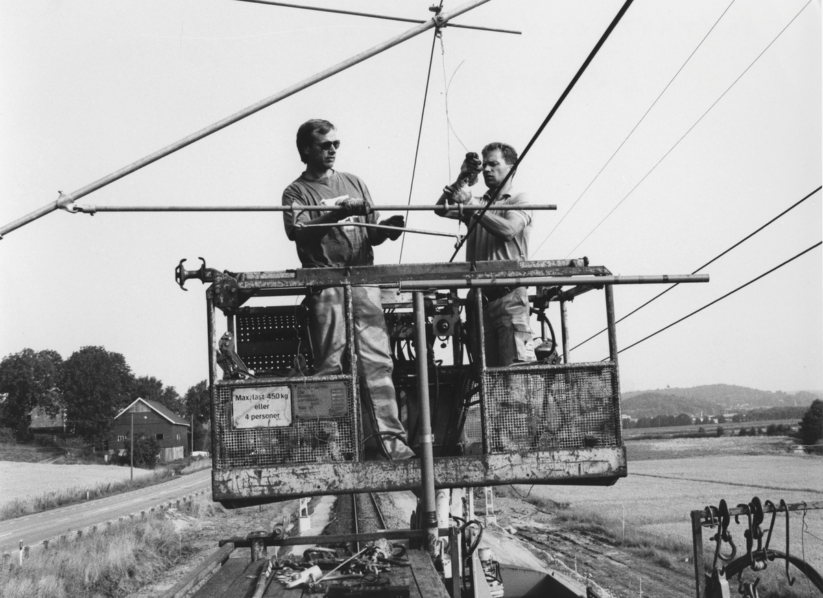Två män monterar kontaktledningar som sker med hjälp av lyftkorgar fastsatta i tåg. De befinner sig över järnvägsspåret i Anneberg, Lindome, hösten 1990-08-31. Relaterat motiv: A3054.