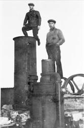 To menn står på en maskin, stort svinghjul bak.