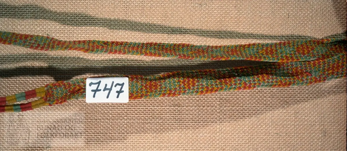 Fingra band av rødt, blågrønt, gult og sauekvitt ullgarn.  I ene enden 2 små tvunnede fletter.  Andre enden 2 fletter med stoppesøm.  L.: 170 cm + 20 cm fletter.  Br.: 2,5 cm.
