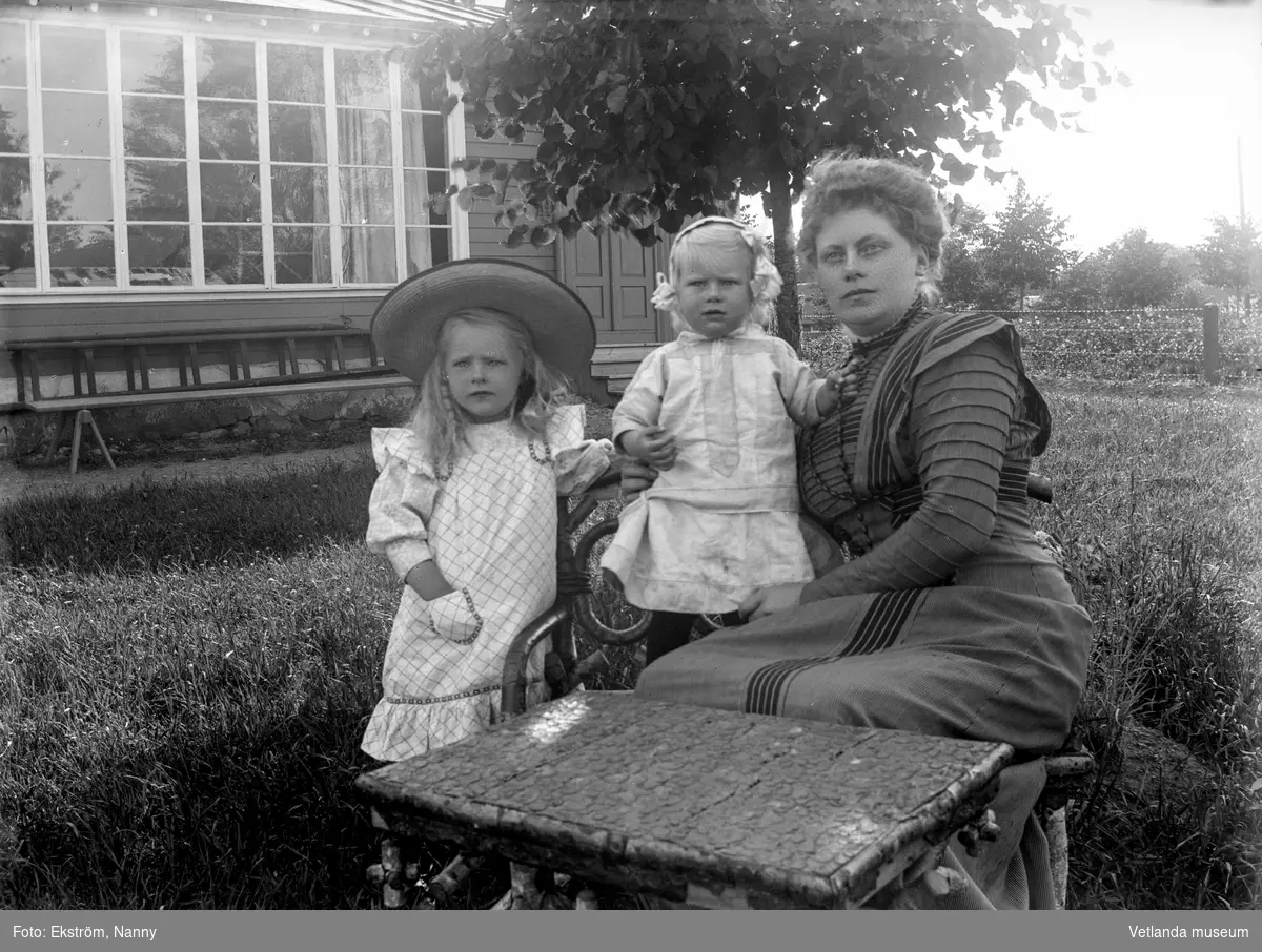 Kvinna med två små barn i trädgården framför fotoateljén på Mogärde i Vetlanda. Kvinnan är troligtvis fotografens syster, Amy Ekström och barnen är troligtvis hennes döttrar, Anna Karin och Inga Kristina.
