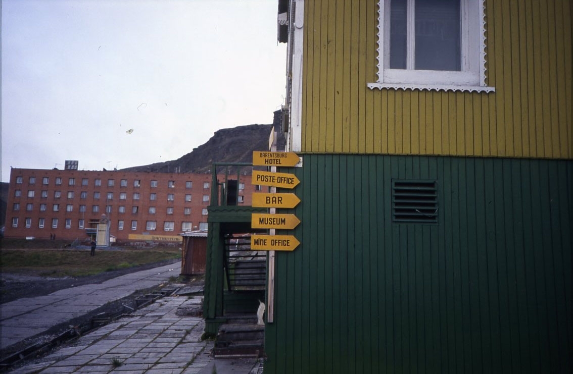 Bebyggelse i det ryska gruvsamhället Barentsburg på Svalbard. En gul hänvisningsskylt i förgrunden som pekar mot Bar, Postkontor, Gruvkontor, Museum och Hotell