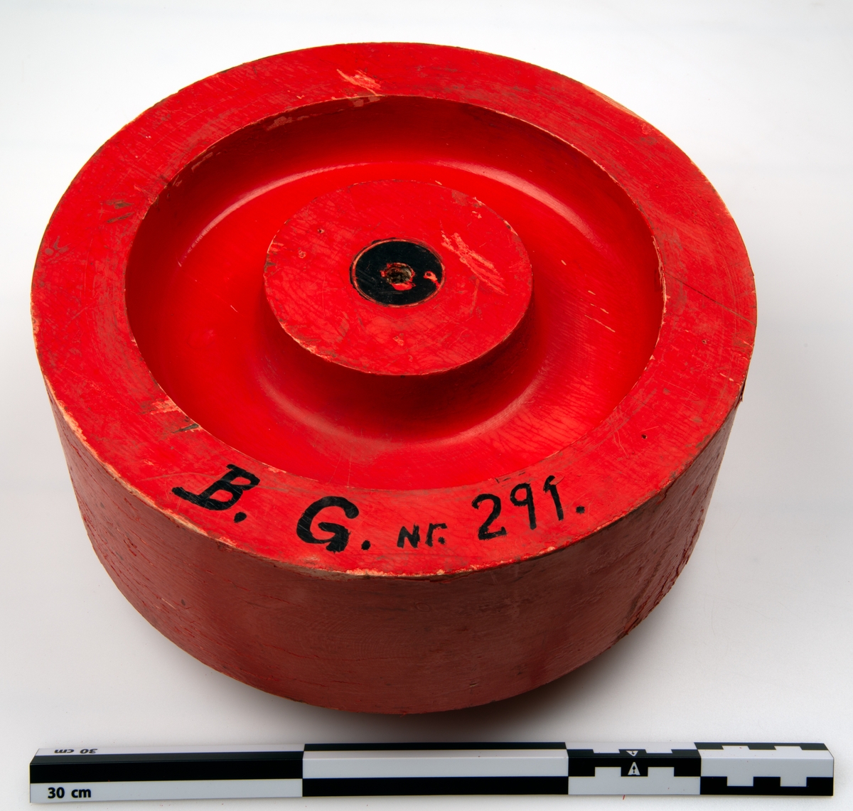 Sirkulær gjenstand, med utsjæringer på en side og utstikkende sylinder på den andre siden. gjenstanden er hovedsalkig malt rød, med detaljer i sort.