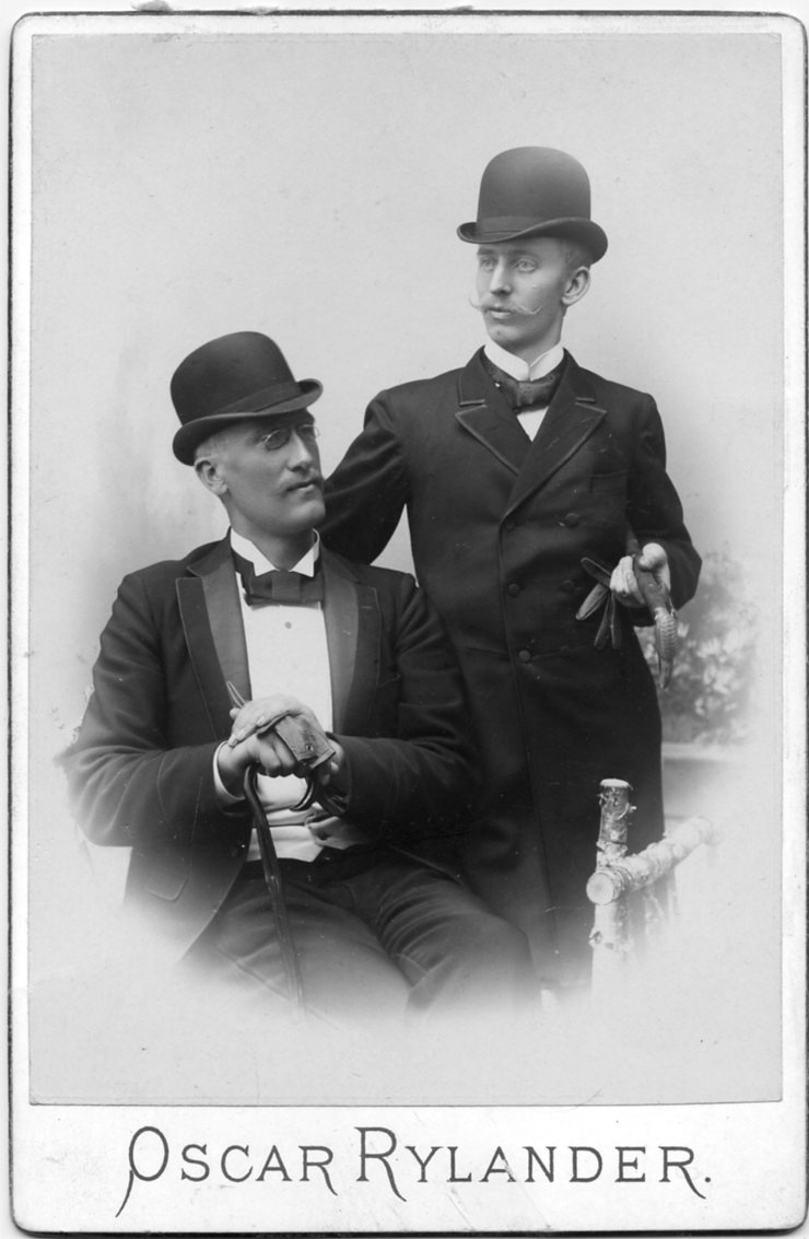 Kabinettsfotografi: två finklädd unga män med kubb, spatserkäpp och handskar. En sitter och en den andre står bredvid.