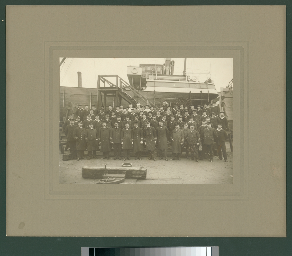 Bilden visar en grupp officerare, underofficerare och manskap från Svenska Flottan som har tagit uppställning för gruppfotografering framför ett träfartyg som ligger på land i ett hamnområde.
