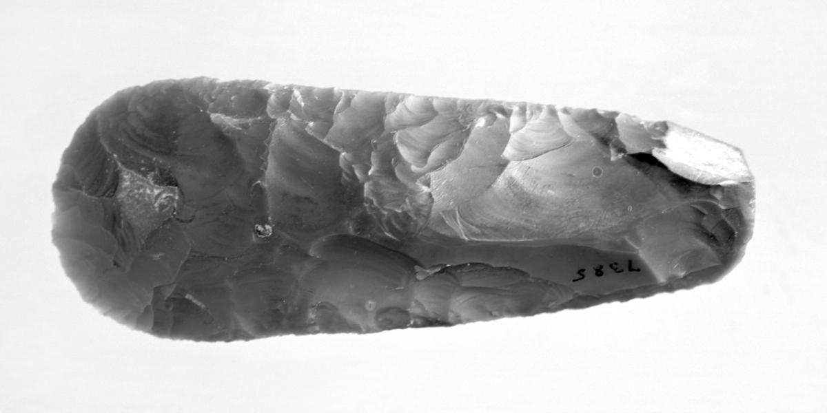 Flintredskap, dannet av en flekke med spor av kalkskorpe. Retusjeret helt rundt undtagen oppe ved slagbulen, og bare paa rygsiden, mens spalteflaten er glat. Stykket ligner enskeformet skraper, men eggen er for svak. Redskapet maa helst forklares som en matkniv (Rogalands Stenalder fig. 194). Længde 11,1 cm. Fundet ved grøftegravning i en myr straks bak husene paa Folkhøgskulen, Halsnøykloster, Eid s., Fjelberg p., Hordaland, g.-nr. 11. Kniven laa i bunden av en grøft, ca. 1 m. dypt. Høiden over havet 20-22 m. Gave fra stortingsmand Knut Markhus. Avb. fig. 4