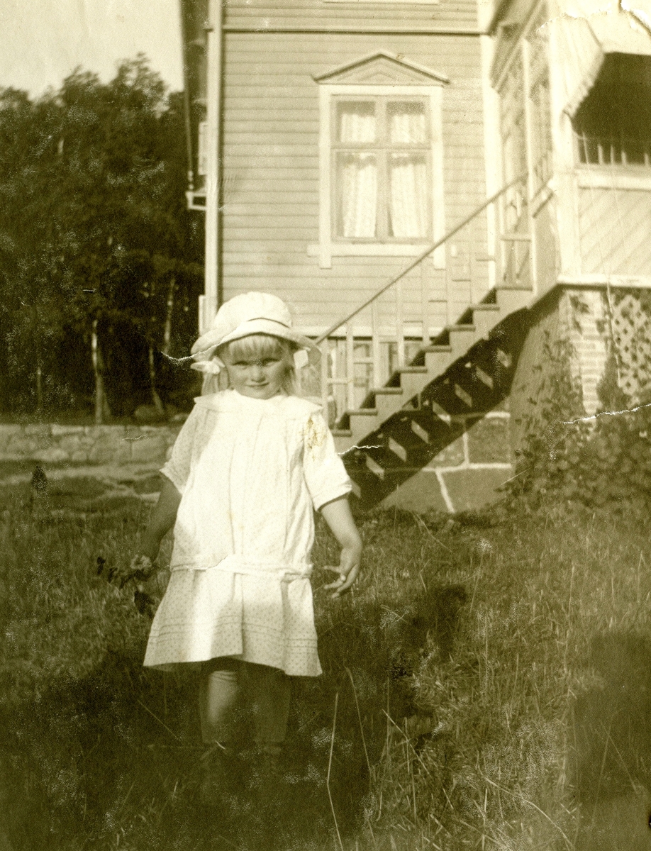 En liten flicka, klädd i kläning och hatt, står i trädgården vid Kållered Stom "Nygård", okänt årtal.