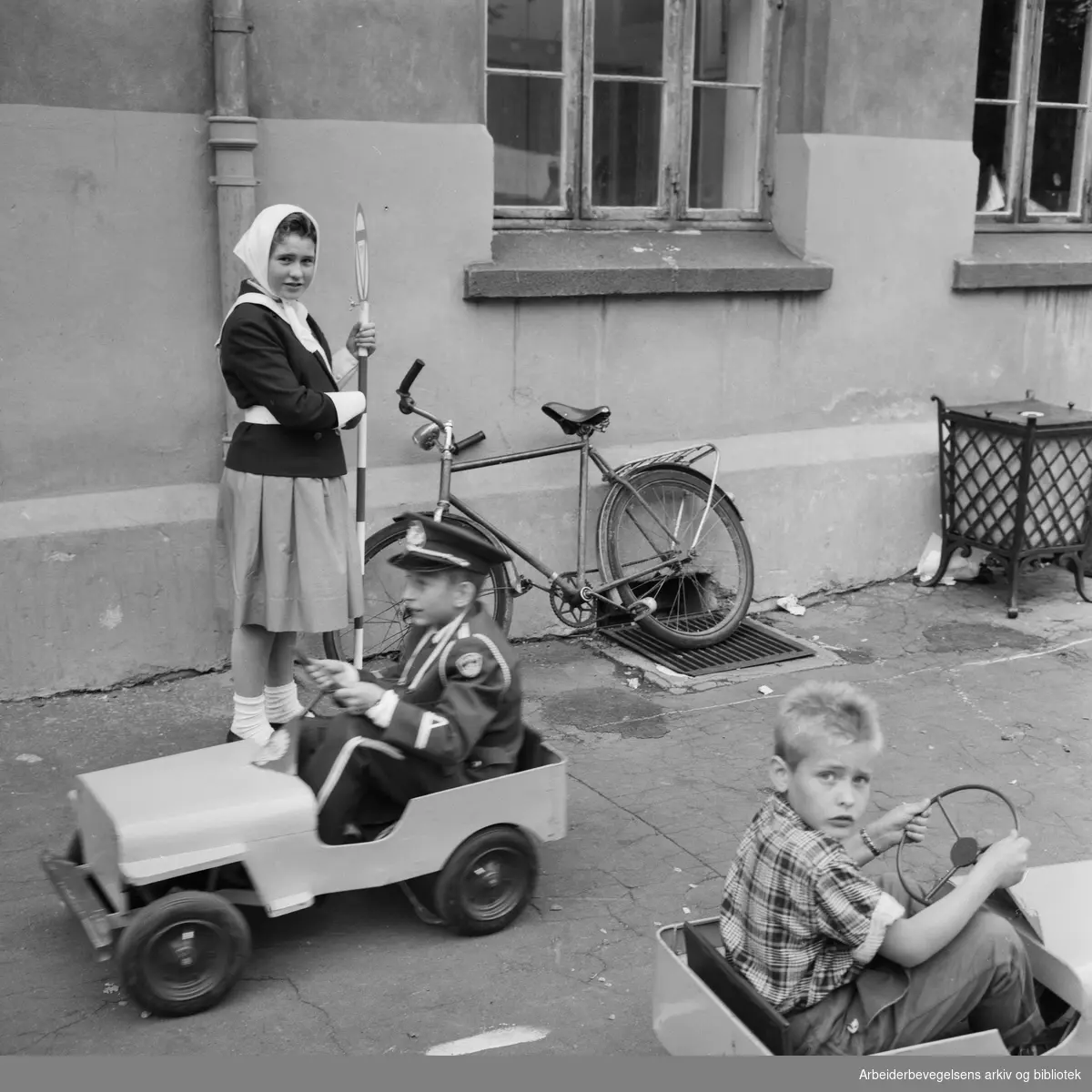 Sagene skole fyller 100 år og arrangerer tråbil-løp i skolegården for elevene. Thor Wilhelm Faye-Hansen i uniform konkurrerer mot Anders Rudlang, mens Kari Randi Fredriksen fra skolepatruljen dirigerer trafikken. Juni 1961.