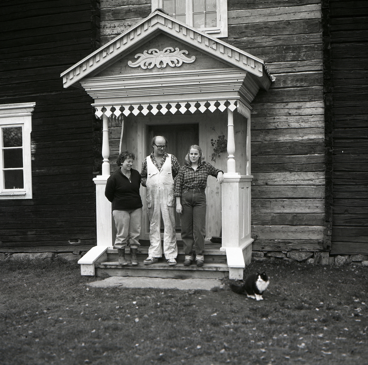Tre personer står uppställda på en förstukvist för fotografering. TV-inspelning om brokvistar på Hymmersbacken i Alfta, 1981. Ett timrat hus renoveras.