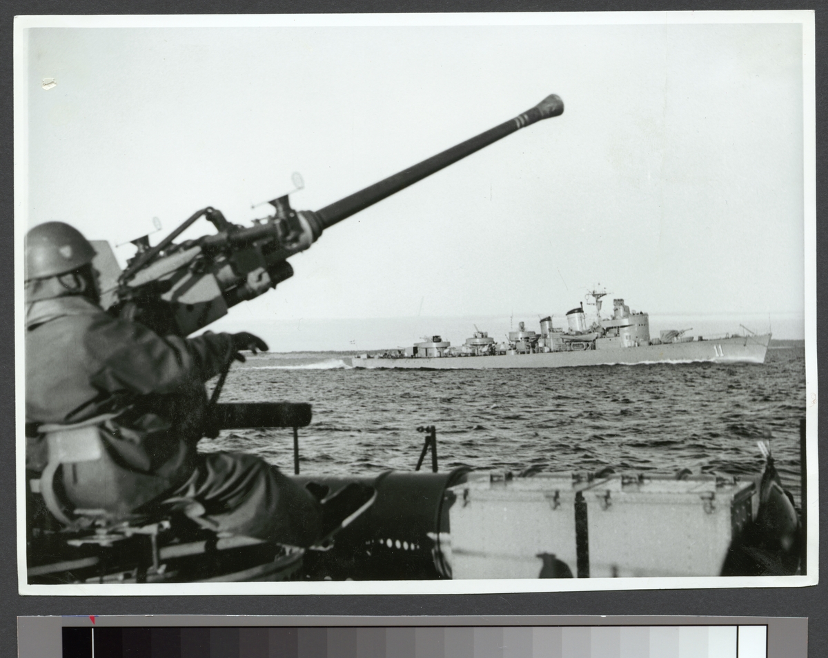 Bilden visar en soldat som siktar med en luftvärnskanon mot himmelen. I bakgrunden syns jagaren Visby.