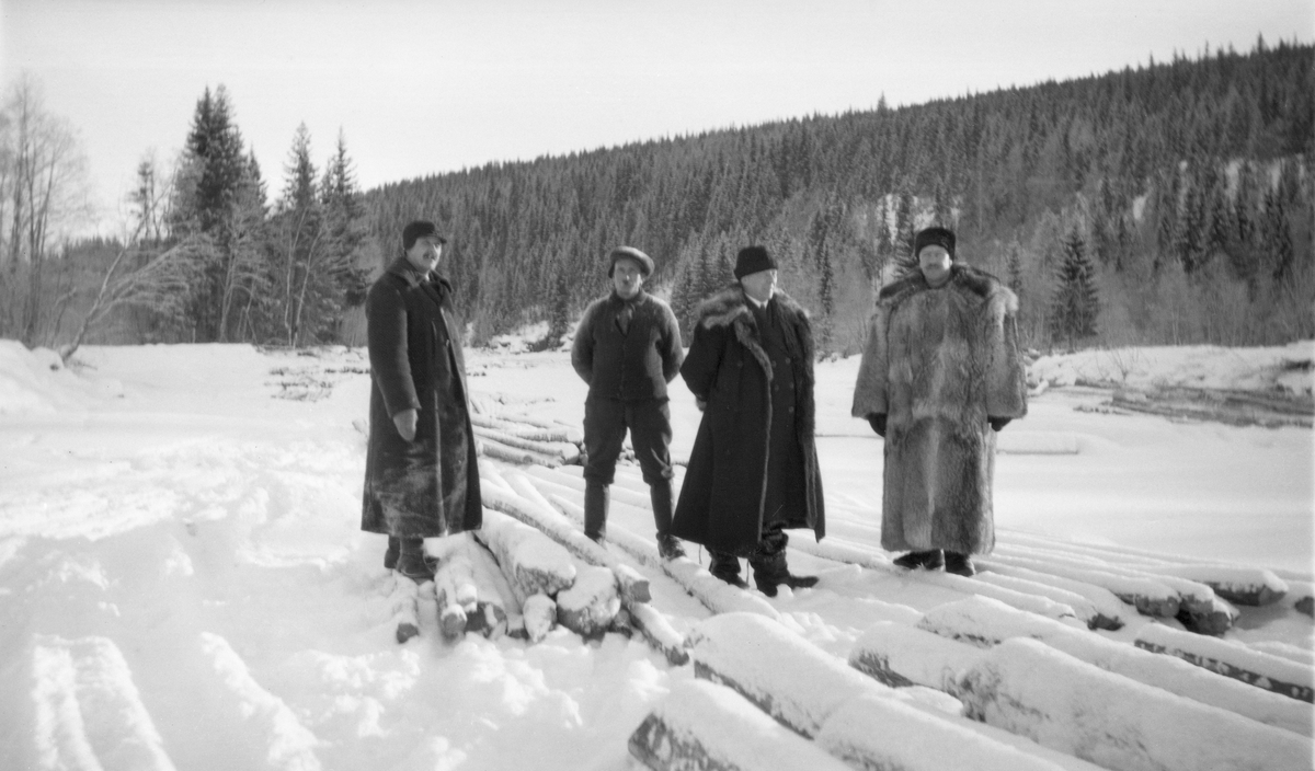 Fire menn, fotografert på isen ved Lermyrberget i den nedre delen av elva Åsta. Bildet skal være tatt i mars 1921. Vi vet dessverre ikke hvem disse mennene var, men et par av dem er også med på fotografiet SJF.1990-01708. Tre av dem var kledd i lange frakker, den ene av ulveskinn. Mannen som sto som nummer to fra venstre var mer arbeidskledd. På isen bak de fire karene ser vi en del tømmer, som antakelig var lagt i såkalte «flakvelter» i påvente av at tømmermålerne skulle komme. Disse målerne var tømmerkjøpernes «annammere» - de skulle besiktige og måle hver enkelt stokk og deretter slå på dem merker, som fortalte hvem som hadde kjøpt stokkene, før våren kom og elvene gikk opp, for da ble jo tømmer til mange kjøpere fløtet nedover det samme vassdraget. Seinere, ved lensene lengre nede i Glomma ble stokkene samlet og sortert etter de nevnte merkene. Flakveltene besto av stokker som var lagt tett og parallelt inntil hverandre i ett lag, vinkelrett på et par underlagsstokker. Dette skulle gjøre det enkelt for tømmermålerne å komme til med måleinstrumentene sine – tømmerklaver – og med merkeøksene. Men da måtte ikke tømmeret være tallagt i flakvelter for tidlig, slik at de ble dekt av store snømengder. Flakveltene krevde dessuten ganske stor plass. Her har man funnet et sted der elva var ganske bred. Det å levere tømmer på is kunne imidlertid skape problemer for fløtinga, så etter hvert kom det påbud om at tømmeret skulle leveres ved, ikke på vassdraget.